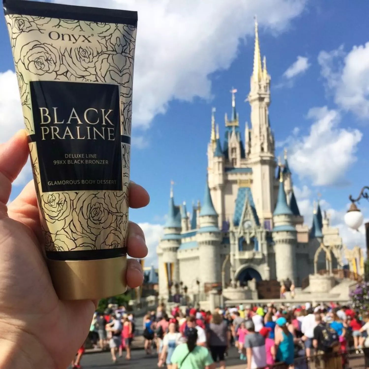 Osoba trzyma tubę balsamu do opalania Onyx Black Praline przed bajkowym zamkiem w słoneczny dzień, w tle widać turystów spacerujących po deptaku.