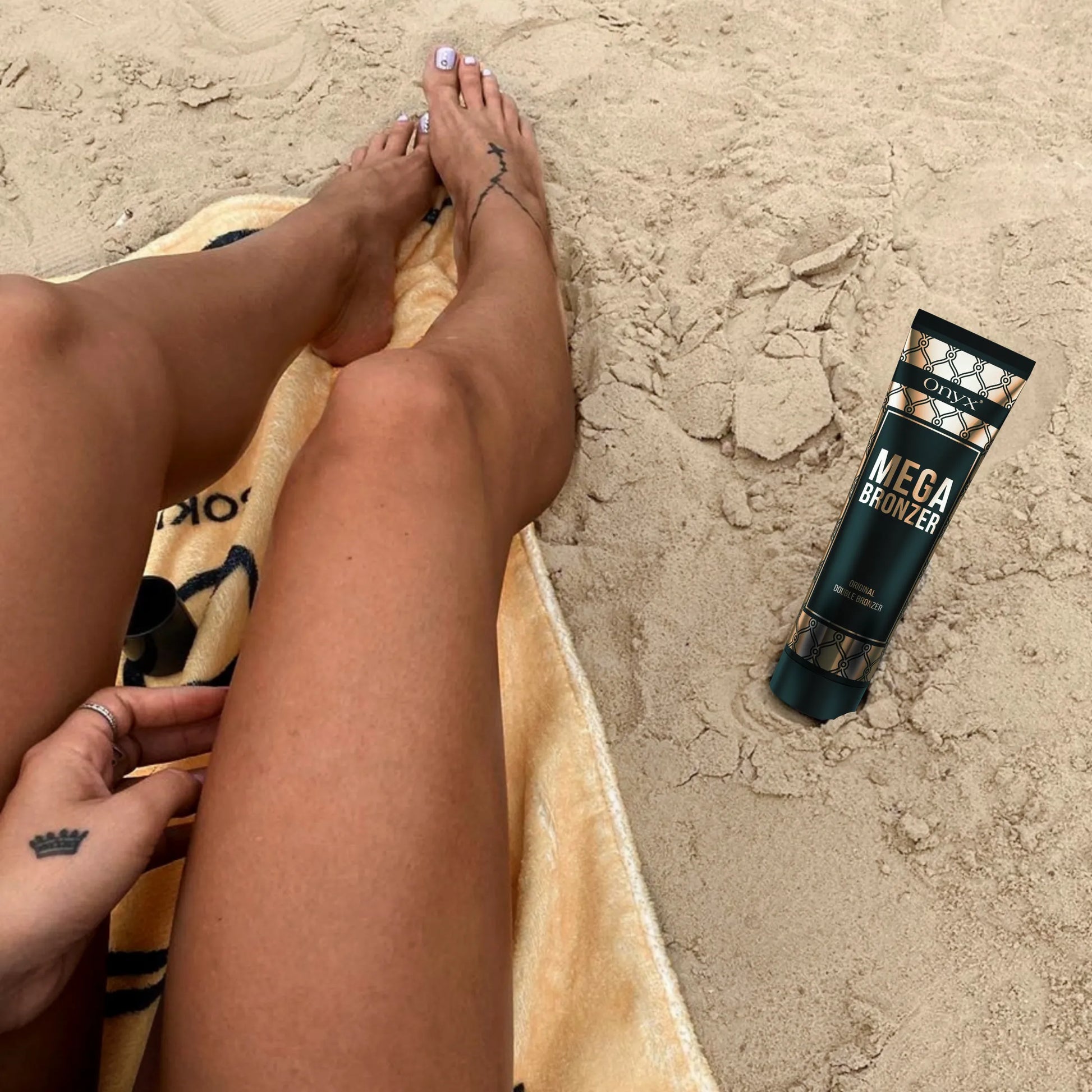 Opalone nogi kobiety leżącej na plaży, obok na piasku balsam brązujący Onyx Mega Bronzer, kosmetyk do podkreślenia koloru skóry na słońcu