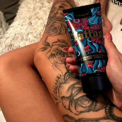 Kobieta trzyma balsam brązujący Onyx Tattoo Bronzer nad tatuażem z motywem róż na nodze, pokazując efekt nawilżonej i rozświetlonej skóry