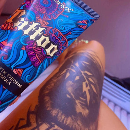 Zbliżenie na tatuaż na udzie i trzymaną tubę balsamu Onyx Tattoo Bronzer w świetle lamp solarium, produkt podkreślający kolor i chroniący tusz przed blaknięciem
