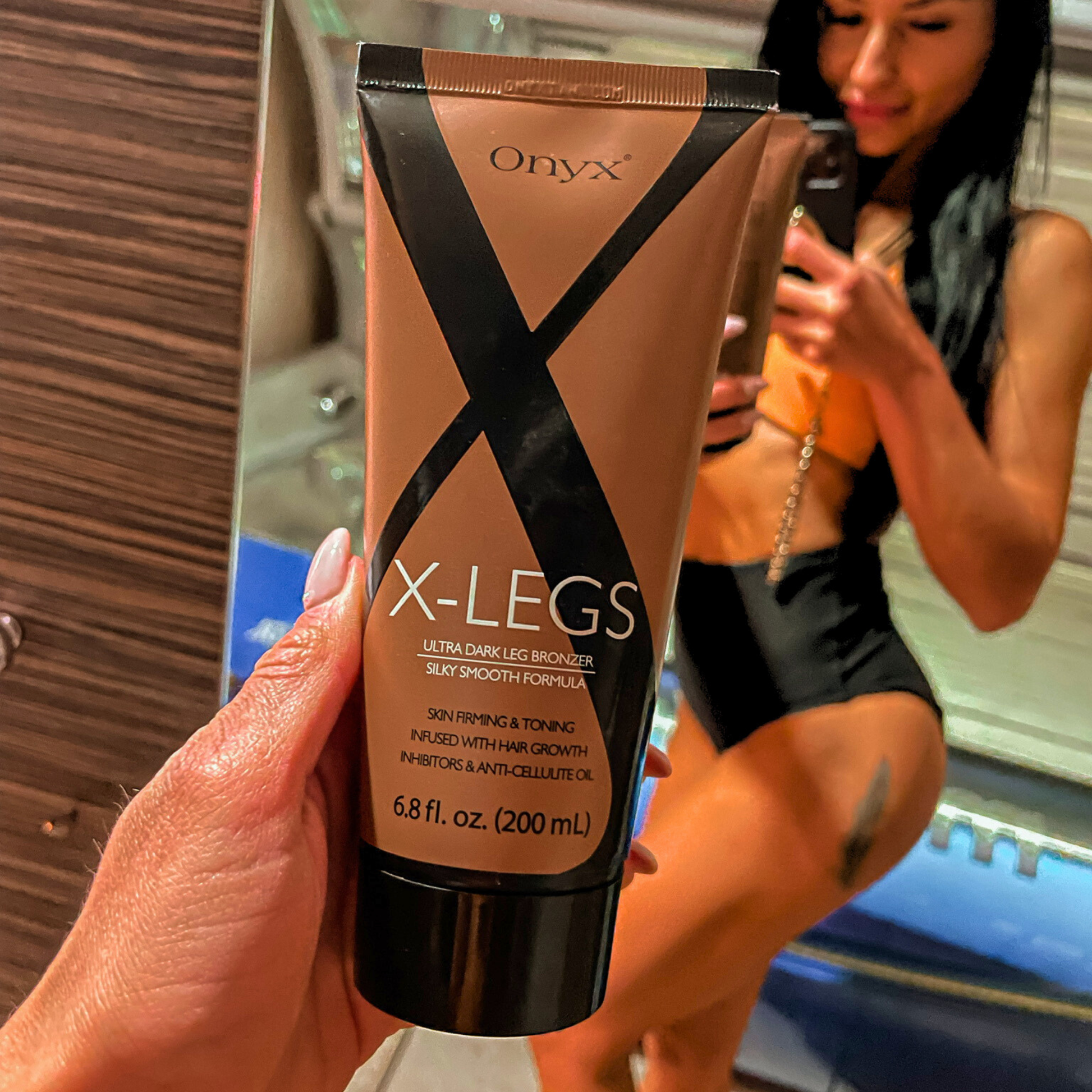 Kobieta z opalonymi nogami trzyma Onyx X-Legs przed lustrem. Bronzer do nóg podkreśla kolor skóry i wspiera jej jędrność.