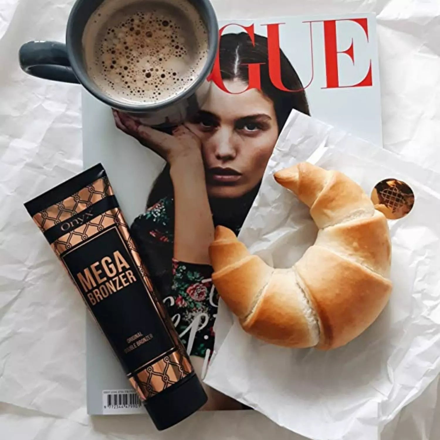 Tuba balsamu do opalania Onyx Mega Bronzer leży na magazynie Vogue obok filiżanki kawy i rogalika, tworząc elegancką kompozycję z produktem do brązującej pielęgnacji skóry